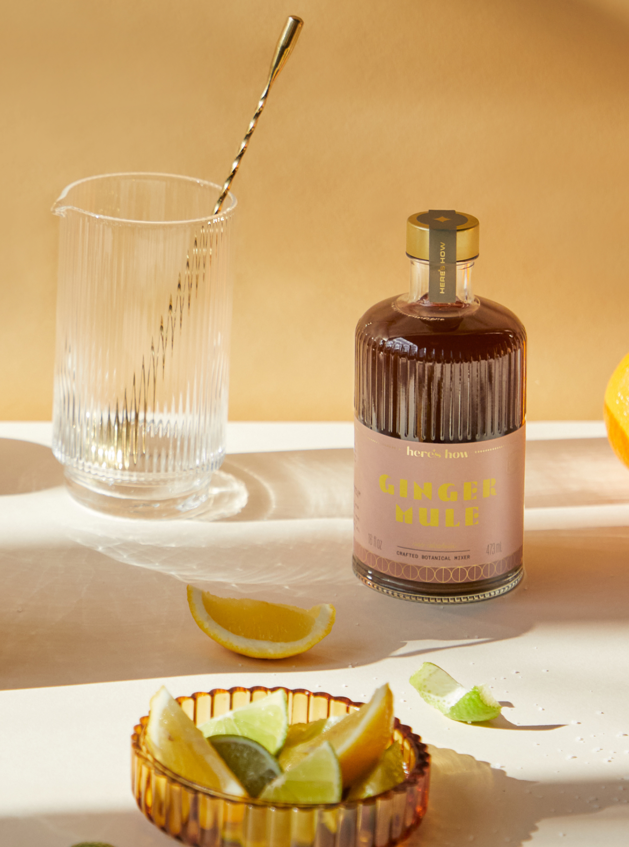 A bottle labeled Ginger Mule sits on a table with a glass pitcher, a metal stirrer, and lemon and lime wedges nearby.