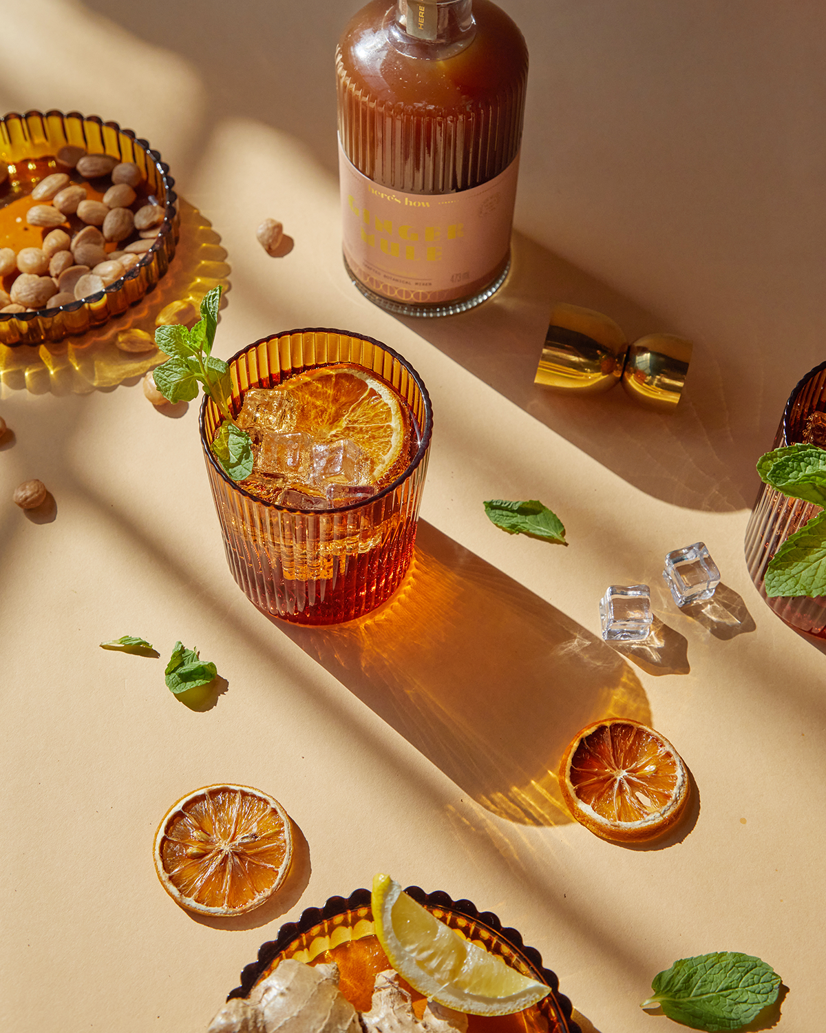A glass of Mookie + Lenny amber soda lime drink with ice and mint sits on a beige surface, surrounded by dried citrus, nuts, and home bar essentials like a bottle and cocktail shaker parts.