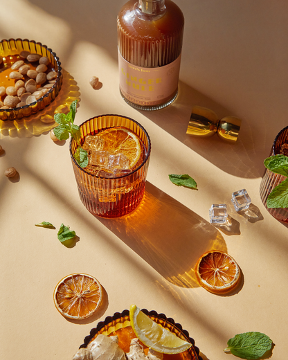 A glass of Mookie + Lenny amber soda lime drink with ice and mint sits on a beige surface, surrounded by dried citrus, nuts, and home bar essentials like a bottle and cocktail shaker parts.