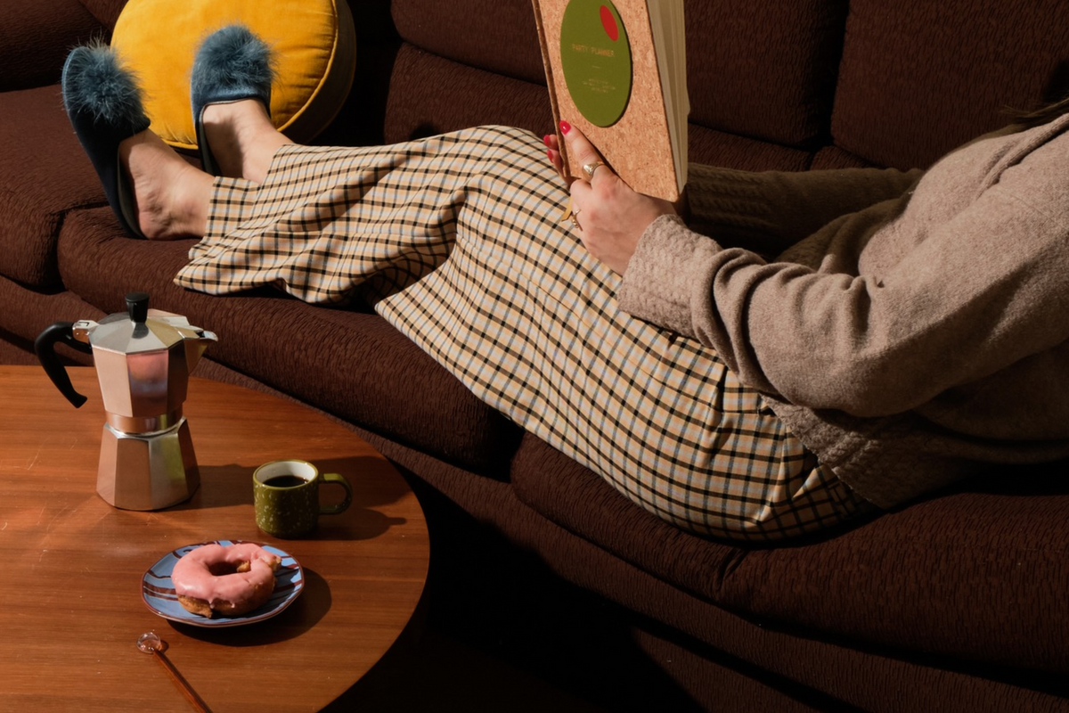 A person reclining on a brown couch with a book in hand, wearing a beige and black checkered skirt and fuzzy black slippers. A wooden coffee table beside them holds a pink donut, a green mug, and a silver moka pot.