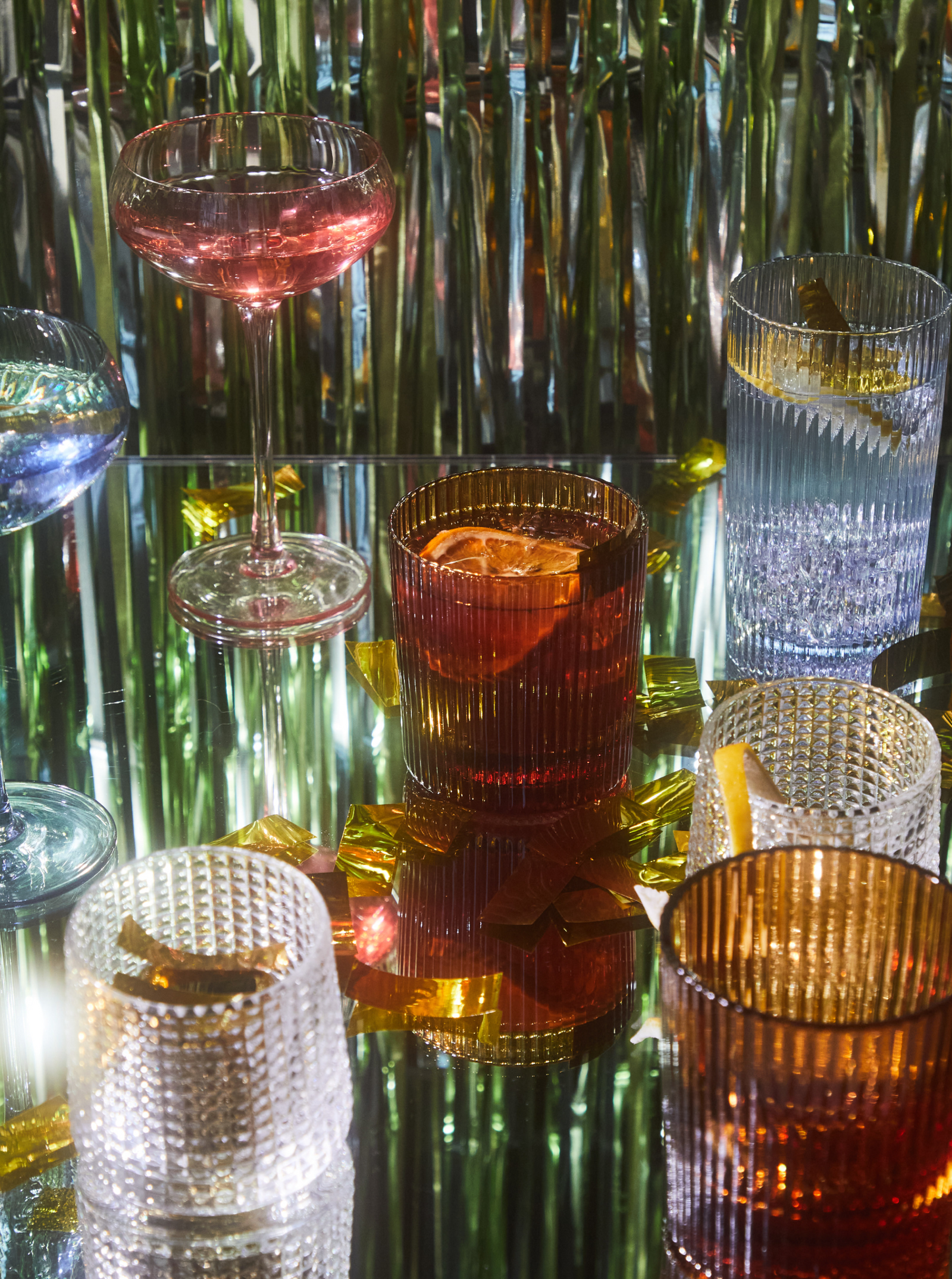 Various colored cocktails with citrus garnishes in textured glasses and a coupe glass, arranged on a reflective surface with metallic and gold ribbon decorations in the background.