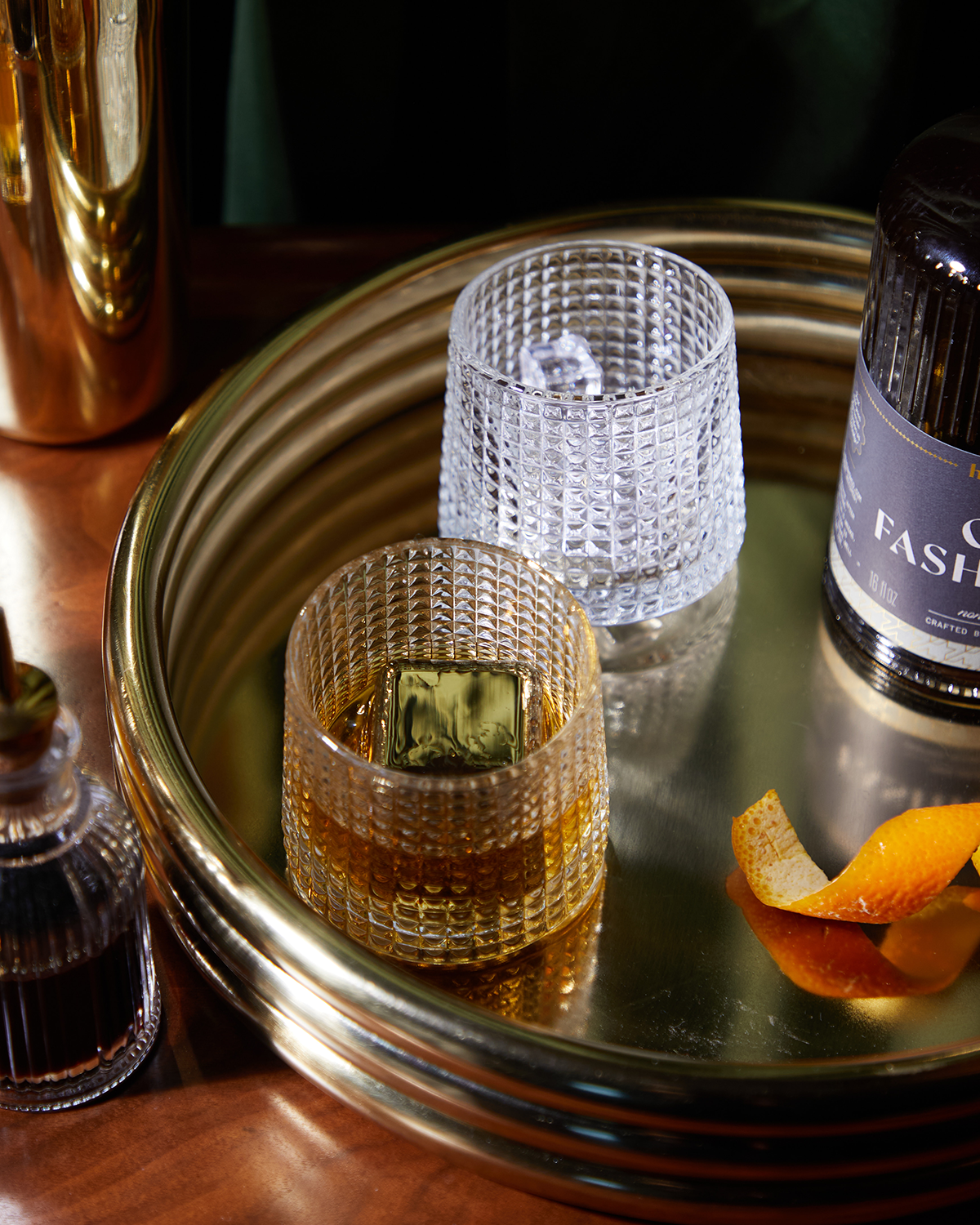 The Jupiter set features two textured glasses—one with ice and clear liquid, the other with amber liquid and stainless-steel chillers—on a gold tray beside a bottle, decanter, and orange peel. Ideal for keeping your drinks cold.