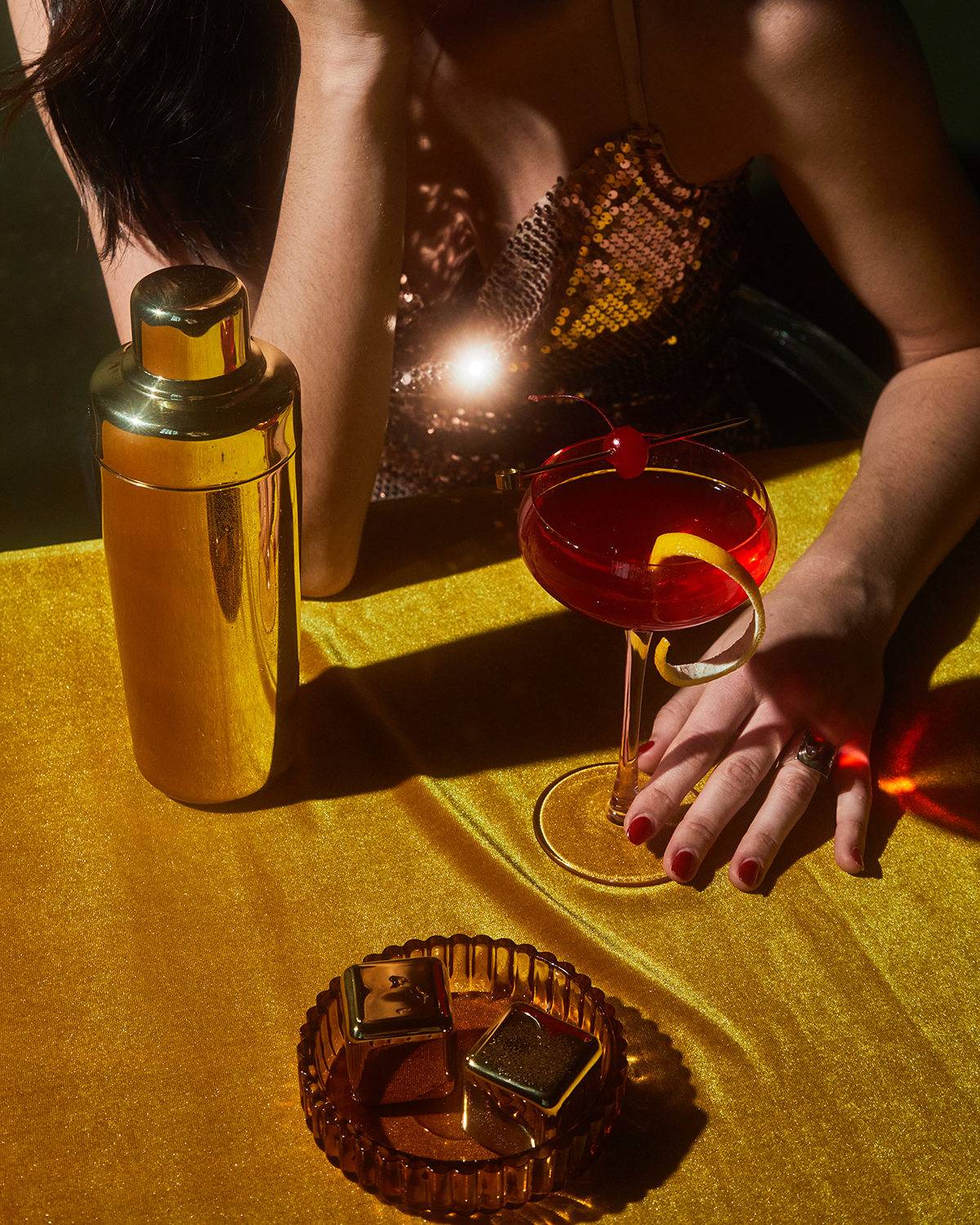 A woman in a sequined top sits at a gold table with The Jupiter cocktail shaker, a red cocktail garnished with a cherry, and an ashtray holding gold-wrapped items. Freezable cubes keep her drink cold in style.