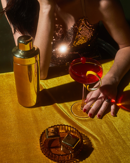 A woman in a sequined top sits at a gold table with The Jupiter cocktail shaker, a red cocktail garnished with a cherry, and an ashtray holding gold-wrapped items. Freezable cubes keep her drink cold in style.