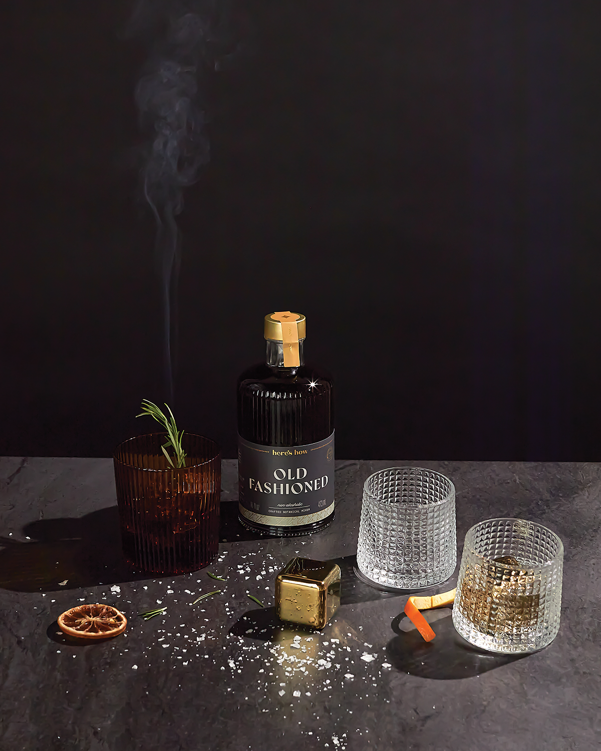 The Jupiter bottle sits on a dark surface with three textured glasses, stainless-steel chillers, a smoking cube, dried orange slice, and rosemary garnish nearby to keep drinks cold in style.