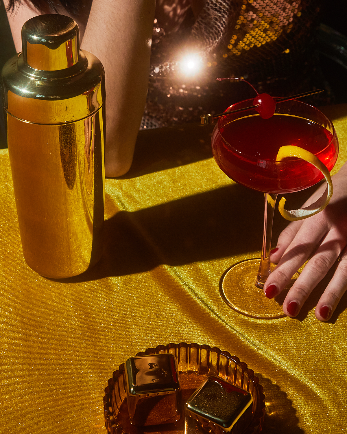 A person with red nail polish holds a cocktail beside The Owen gold shaker and gold-plated cocktail picks on a golden table, with chocolates and sequined clothing in view.